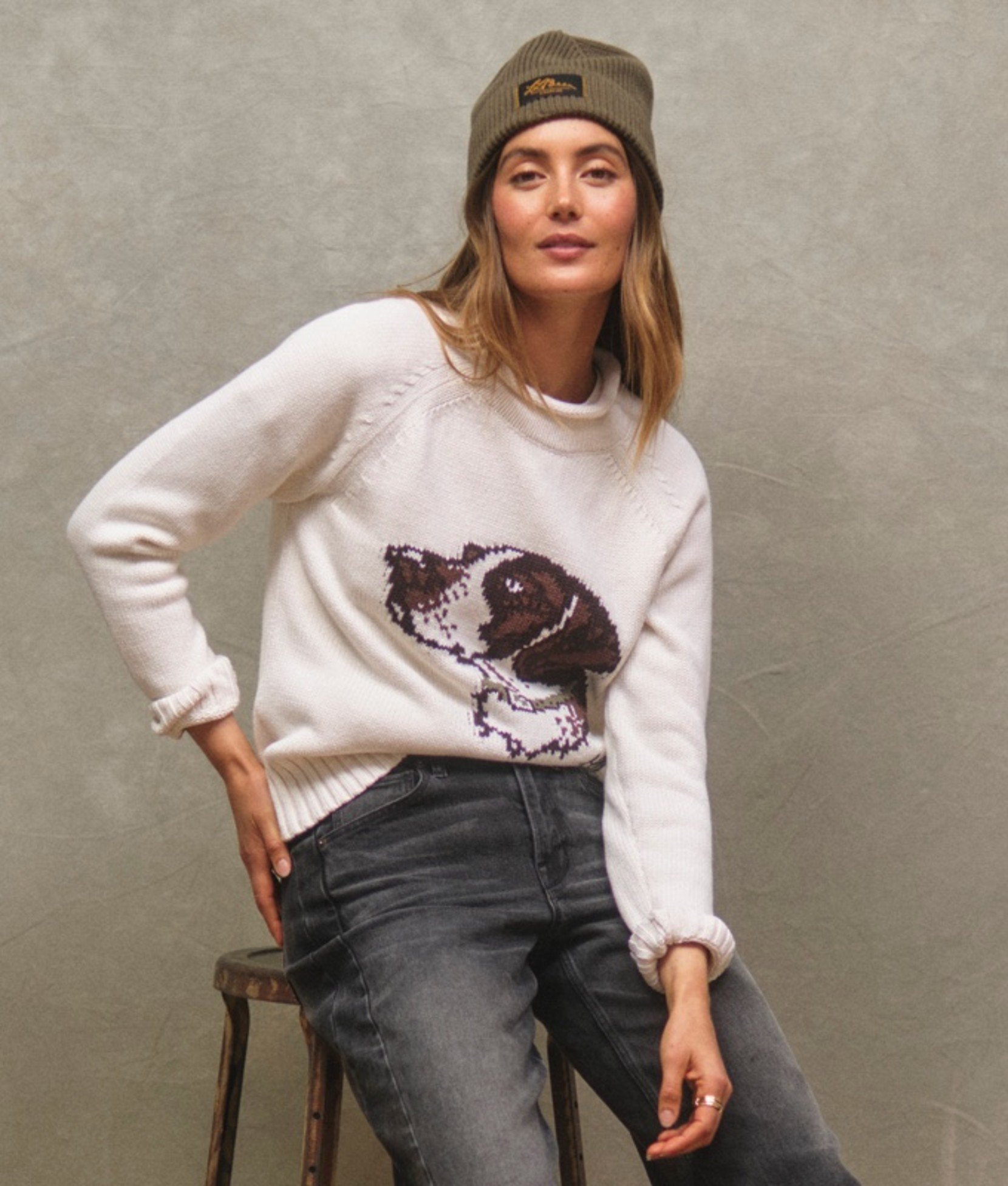 A woman in white dog-print sweater, gray jeans, and olive beanie. 