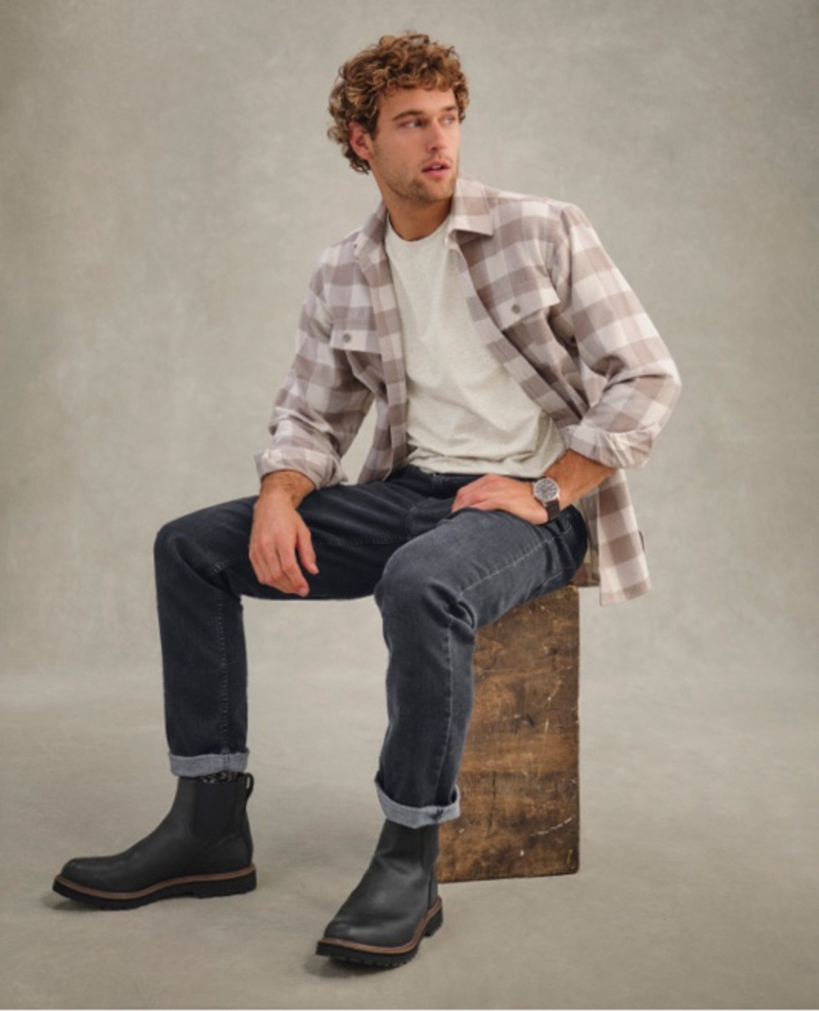 Person in plaid shirt, white tee, jeans, and boots sits on wooden block.