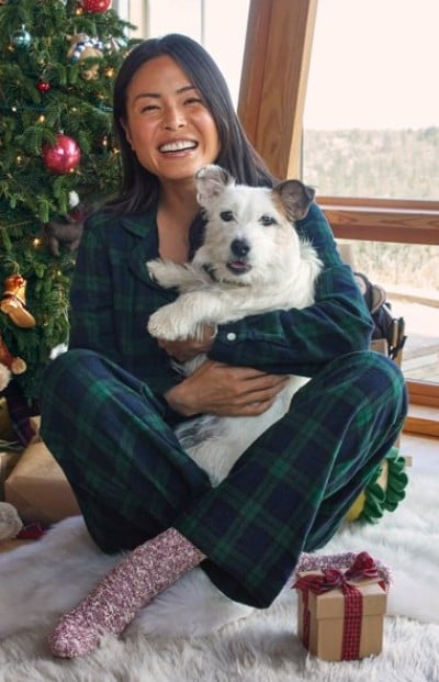 Person in plaid pajamas sits on white rug holding a dog, with a Christmas tree and wrapped gift nearby.