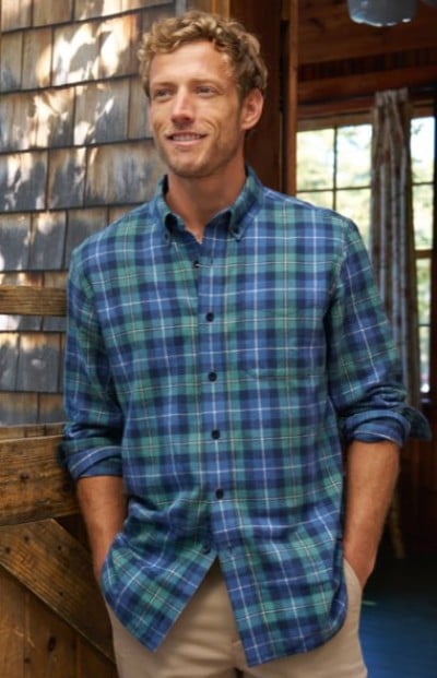 A person in a blue and green plaid button-down shirt paired with tan pants, styled in a rustic outdoor setting.