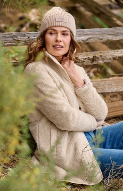 Woman wearing a beige fleece jacket, blue jeans and beanie sits outdoors against a wooden fence, surrounded by greenery. 
