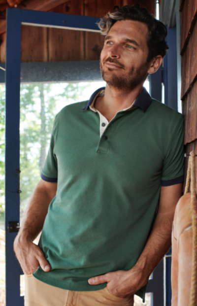 A man wearing a green polo shirt and beige pants with hands in pockets leans against a cabin door.