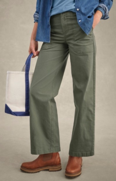 Cropped image of a woman in blue shirt, green pants, and brown shoes holds a white tote bag with blue handles.