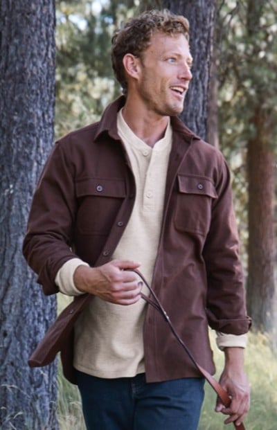 Man in brown jacket and beige shirt walks outdoors holding a leash, with trees in the background.