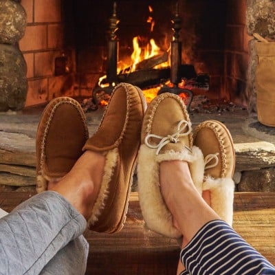 Two pairs of shearling-lined moccasin slippers resting by a stone fireplace with a glowing fire.