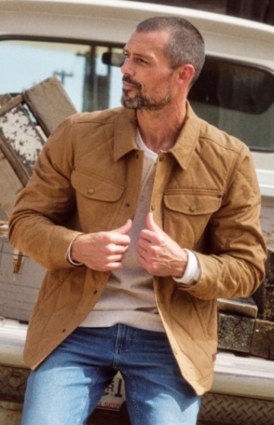 Person sitting on a truck tailgate outdoors wearing layers under a tan jacket and jeans.