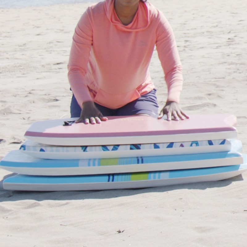 Stephanie kneeling in front of a stack of 4 boogie boards on the sand.