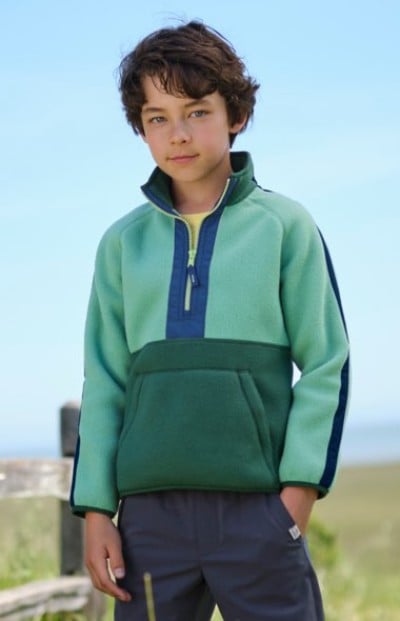 A child standing outdoors wearing a green colorblock pullover with one hand in pocket.
