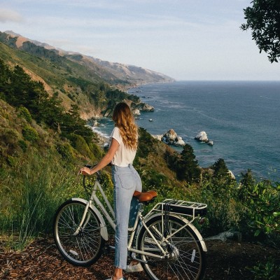 A woman straddling a bike on a high path looking out at the coastline and ocean below.