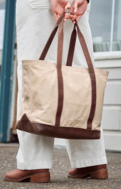 Person carrying a cream canvas tote bag with brown leather trim near a doorway.