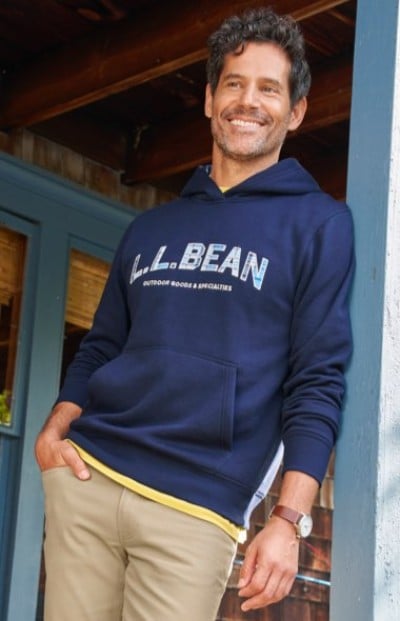 Man in a navy L. L. Bean hoodie and khaki pants leans casually against a doorway outside a wooden building.