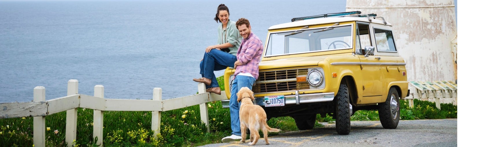 Two people sit by a coastal fence beside a yellow vintage SUV, with a dog standing nearby and ocean cliffs in the background.