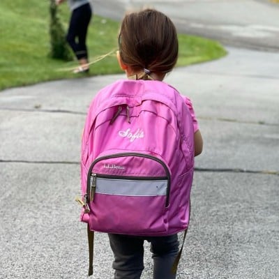 Close-up from behind of a young girl wearing a pink, monogrammed school backpack.