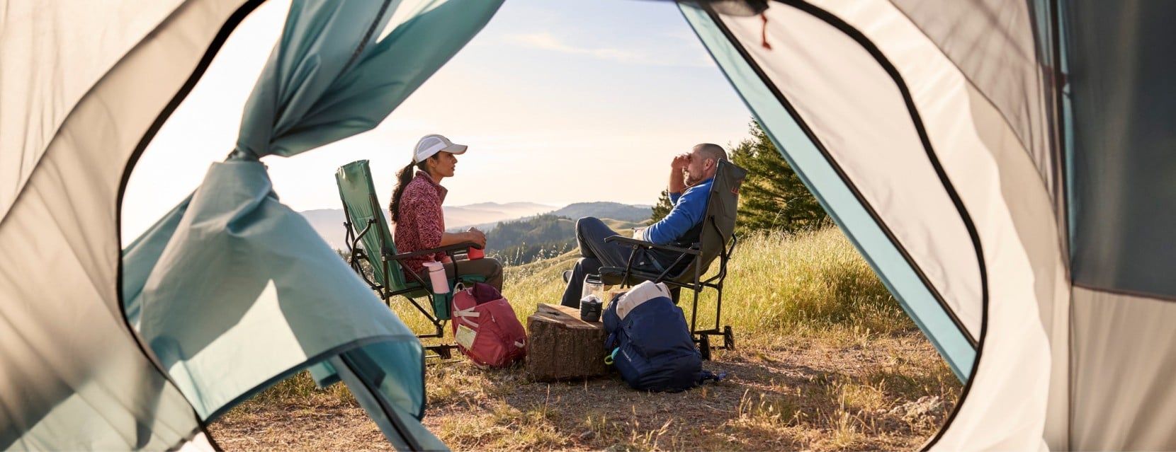 View from inside a tent of two campers seated in chairs, overlooking rolling hills at sunrise with gear nearby.