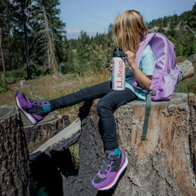 A young girl sitting outside on a tree stump,drinking from an L. L. Bean water bottle and wearing a school backpack.