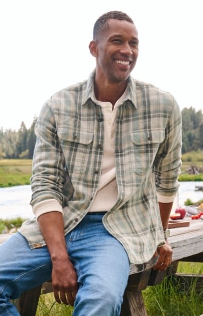 Man in plaid shirt and jeans sits on a wooden bench outdoors, smiling with a river and trees in the background.