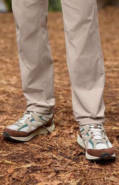 A person wearing beige pants and white, green, and brown hiking shoes stands on a forest floor covered with dried leaves.