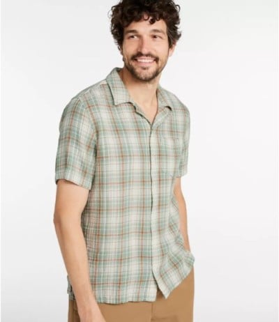 Person wearing a green plaid short-sleeve shirt and tan pants, standing against a plain light gray background, looking to the side.