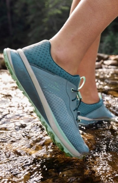 Woman wearing blue running shoes standing in a shallow stream with water flowing over rocks.