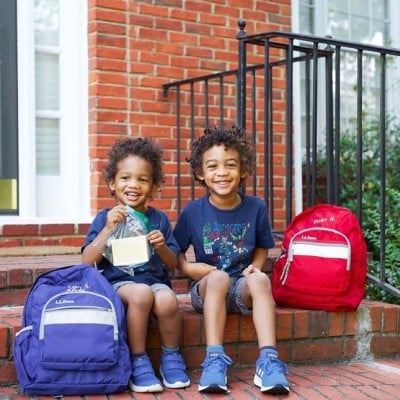 2 kids sitting outside on a front step on the first day of school, L. L. Bean school bacpacks on the ground beside them.