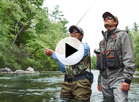 Two people in the river wearing fishing gear, while one is fly fishing.