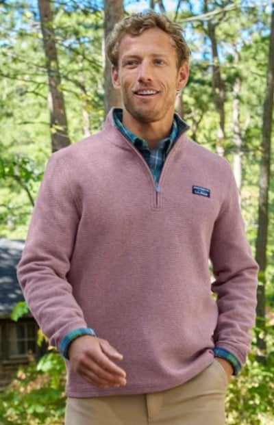 A person in a light pink quarter-zip pullover layered over a blue-green plaid shirt, standing in a forest setting.