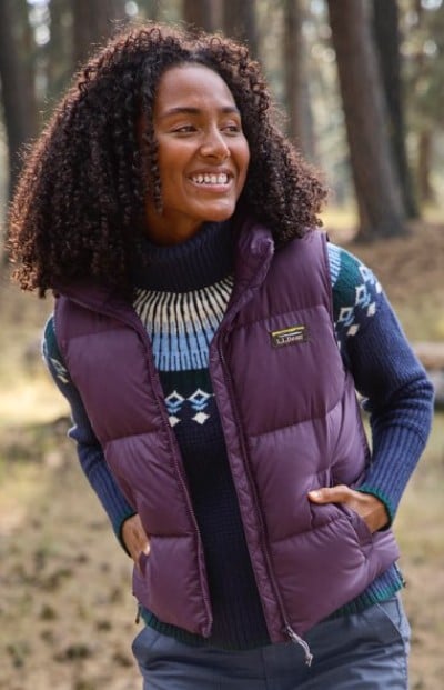 A person in a purple puffer vest and blue patterned sweater walks outdoors in a forested area.