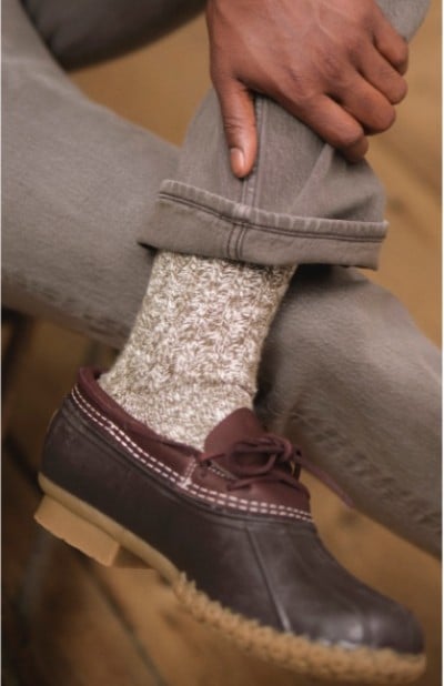 Close up of a person sitting with legs crossed wearing gray pants, gray wool socks, and brown L. L. Bean rubber mocs.