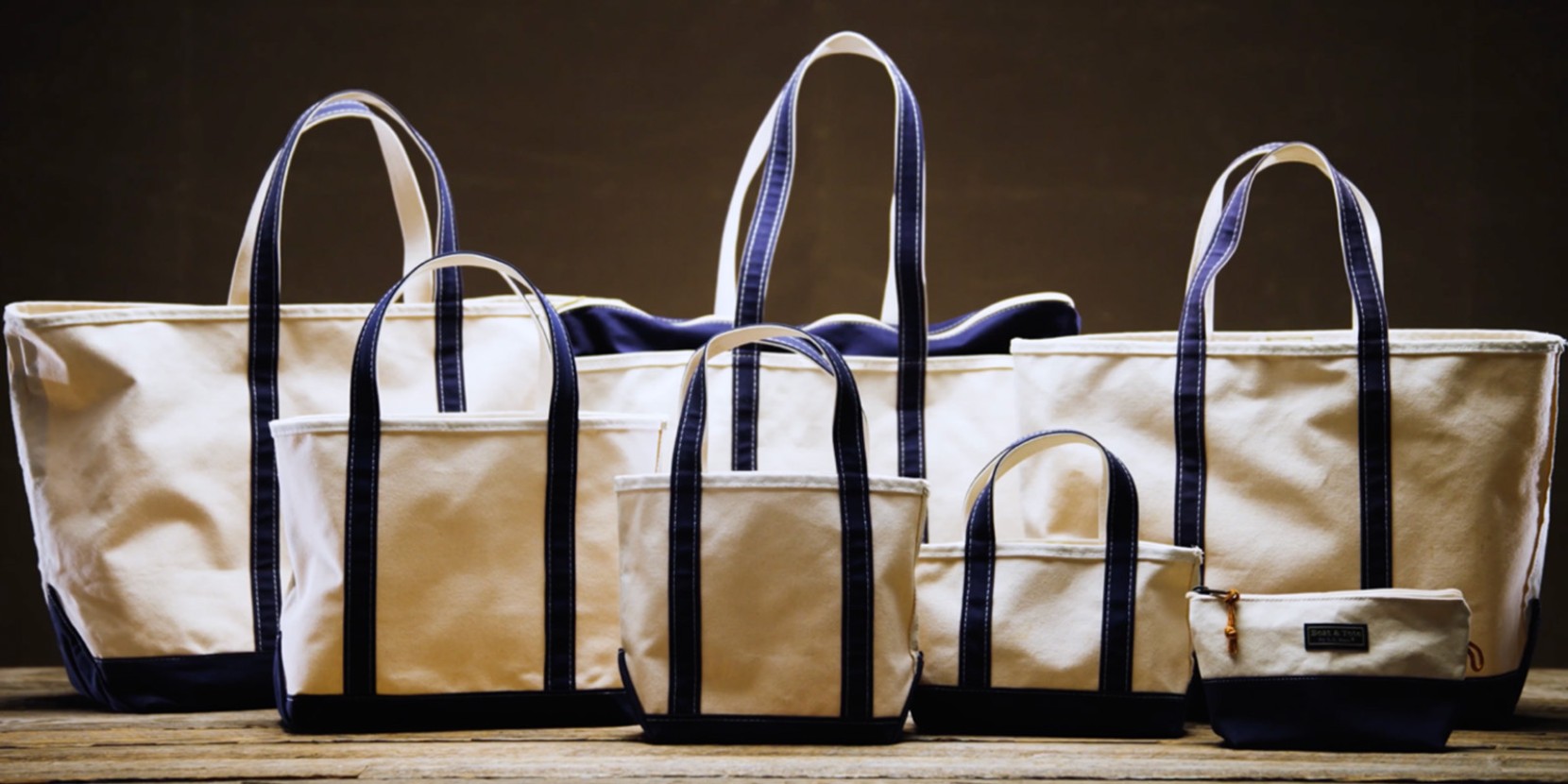 Varying sized white canvas tote bags with dark blue handles and trim, lined up on a wooden surface.