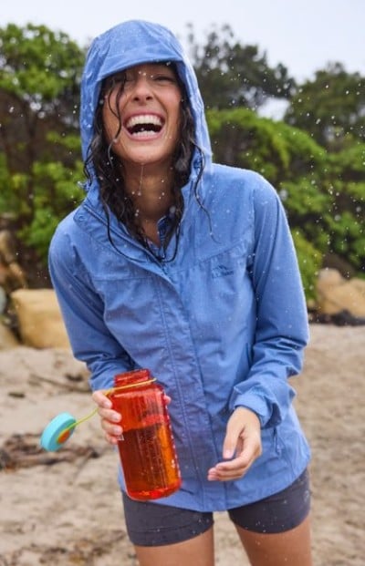 Person in a blue rain jacket on a beach holds an orange water bottle as water splashes around them.