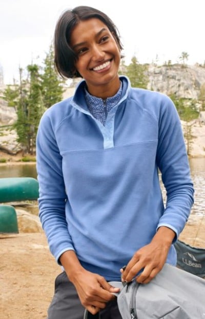 Person wearing a light blue pullover and patterned shirt,stands near a lake with trees and canoes in the background.