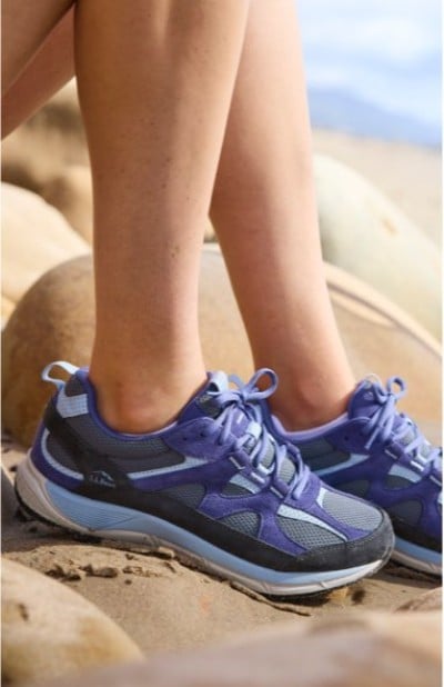 Close up of blue and black athletic shoes on rocky terrain with legs visible against a bright outdoor backdrop.