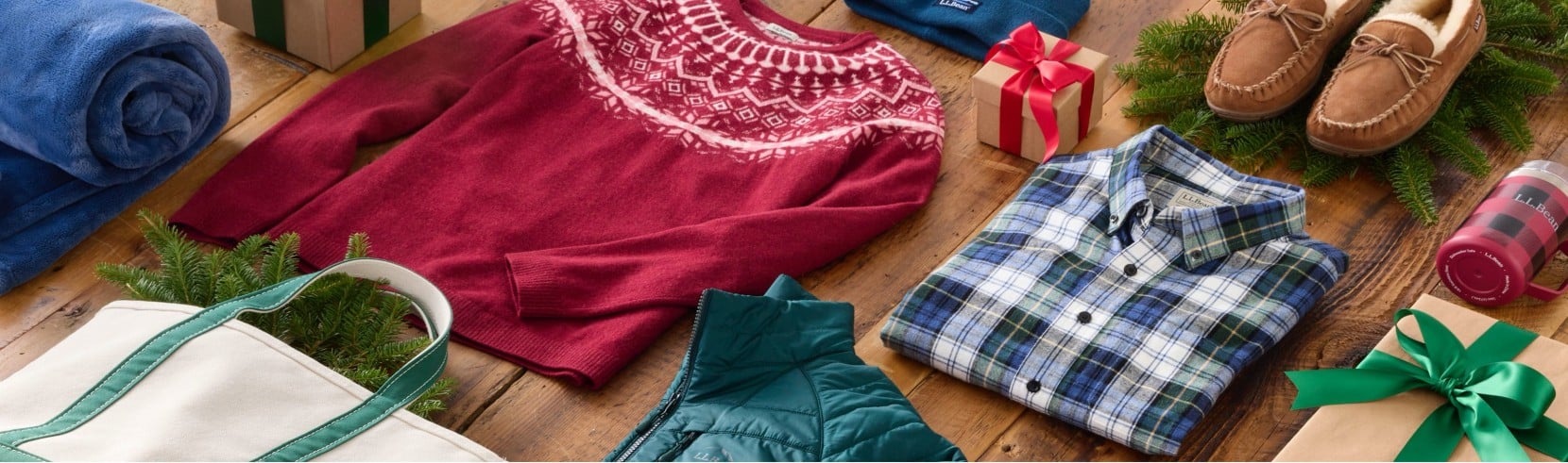Winter clothes, wrapped gifts, a white tote bag, and pine branches arranged on a wooden floor in a festive holiday display.