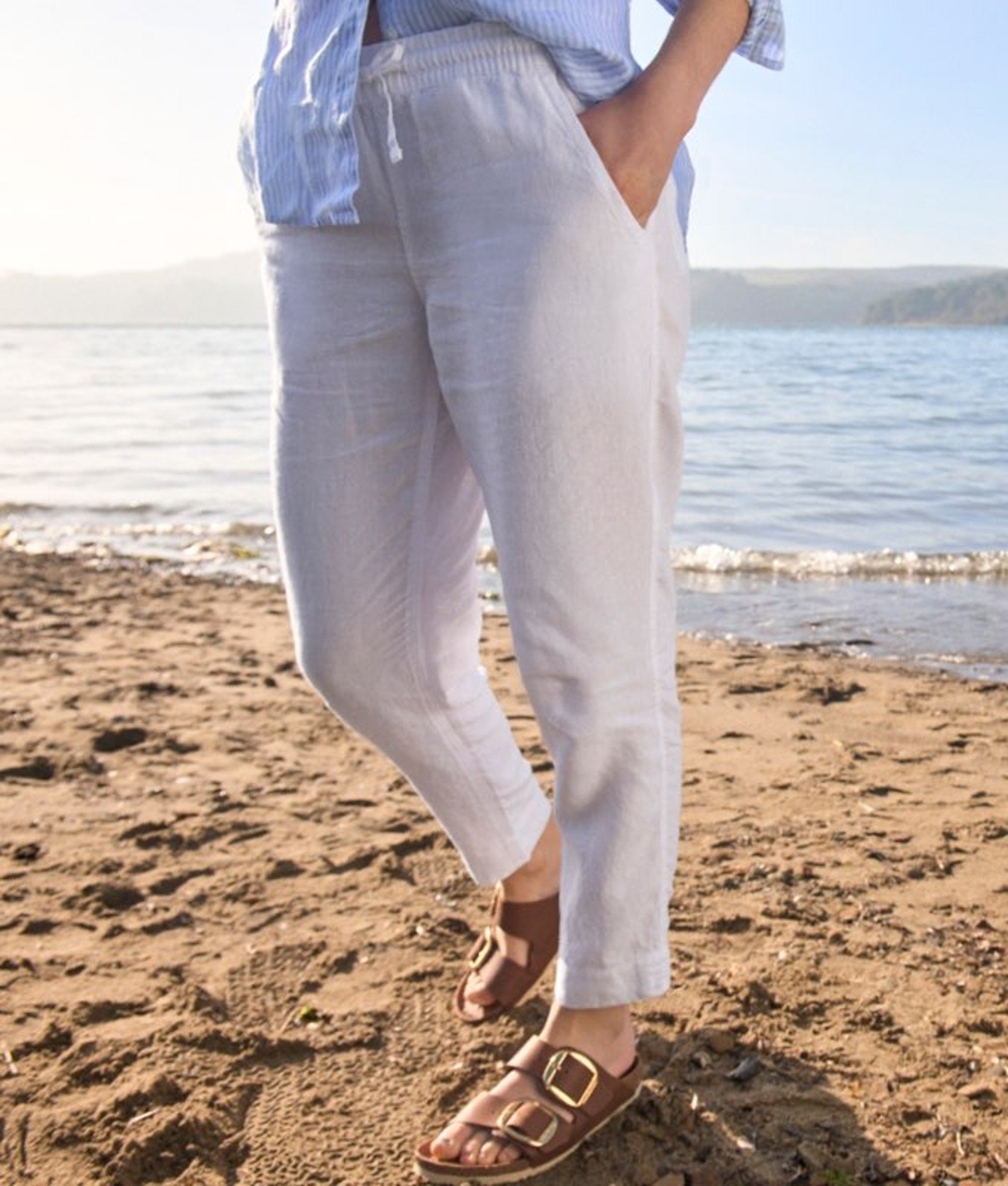 Person standing on sand wearing light-colored pants, with waves and shoreline in the background.