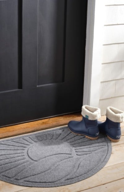 Blue winter boots with white fur lining sit on a wooden porch floor beside a black door and gray semicircular doormat.