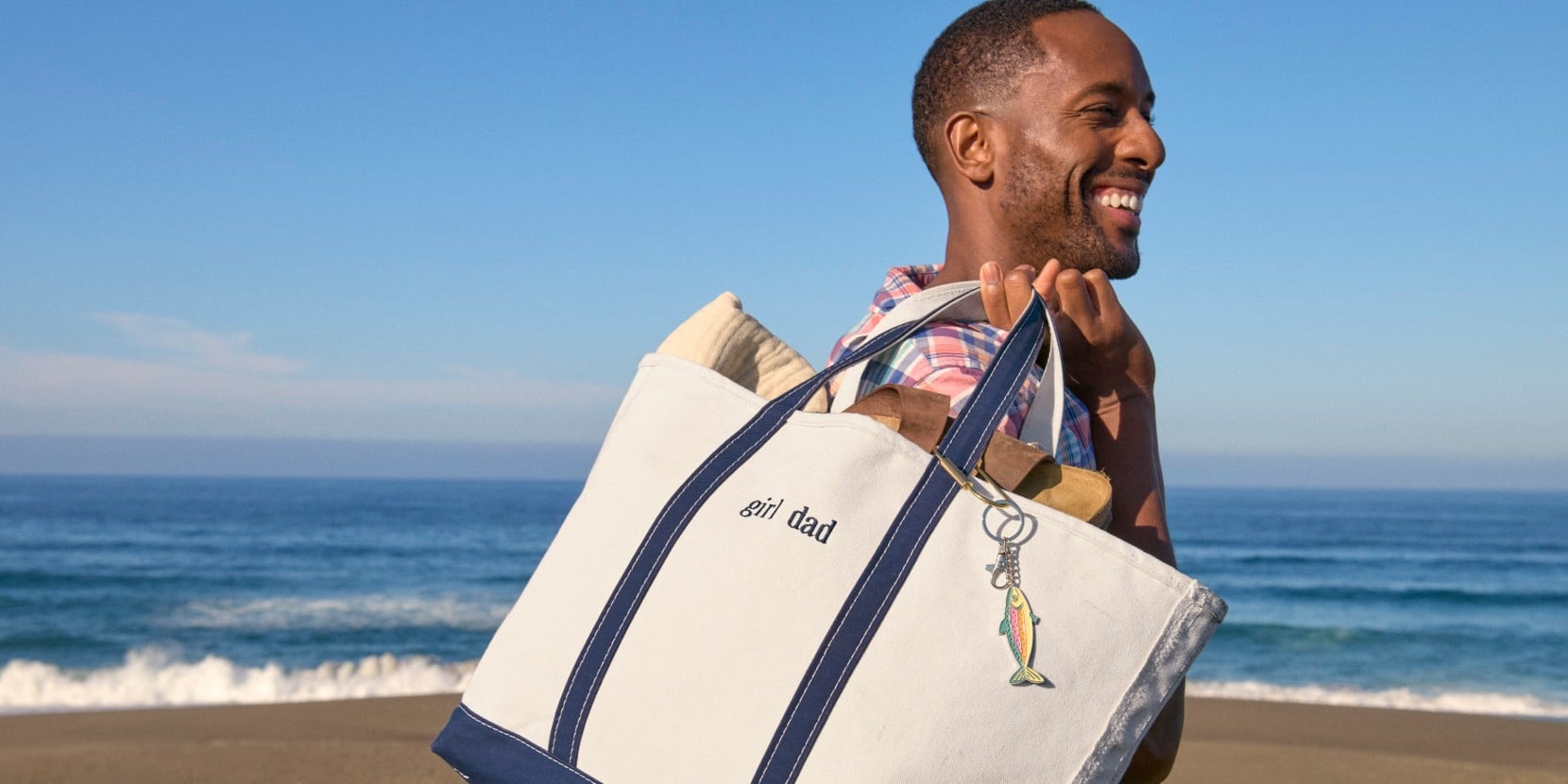 Person walks along a beach carrying a canvas tote with navy straps and keychain, ocean waves and blue sky behind. 