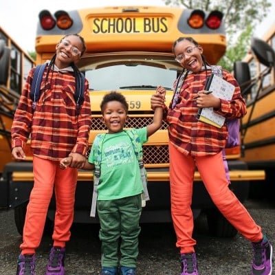 3 kids in front of a yellow school bus wearing school backpacks.