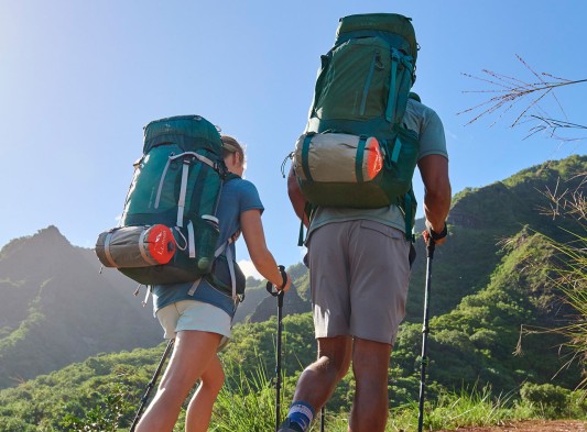 Two people wearing hiking packs, hiking up a trail with mountain in the background.